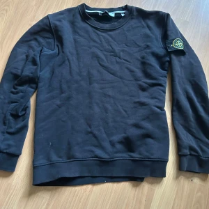 Svart sweatshirt från Stone Island - Säljer en svart sweatshirt från Stone Island med klassisk rund hals och den ikoniska loggan på vänster ärm. Tröjan har långa ärmar och är tillverkad i mjukt bomullsmaterial. Perfekt för en stilren och avslappnad look.