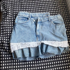 Jeanskjol med spetsvolang från H&M - Supersöt ljusblå jeanskjol från H&M med volangkant i vit broderad spets längst ner. Klassisk femficksmodell med bälteshällor och dragkedja fram. Perfekt för dig som gillar denim med en extra touch av romantisk stil.