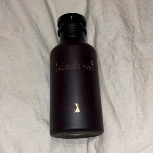 Soleil d'Ombre Jacques Yves parfym - En lyxig parfymflaska i matt mörklila glas med guldfärgade detaljer och svart lock. Flaskan har en elegant och stilren design med texten 'Soleil d'Ombre' och 'Jacques Yves' i guld. Perfekt för dig som gillar exklusiva dofter och snygg inredning på hyllan.
