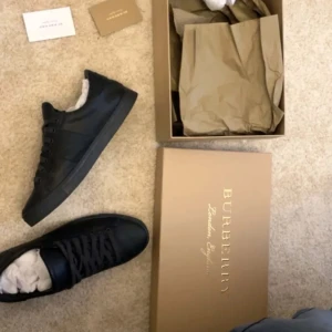 Burberry sneakers - Stilrena svarta sneakers från Burberry med diskret rutig panel på sidorna och präglad logga på hälen. Skorna har rund tå, platt sula och är tillverkade i skinn och textil. Klassisk snörning och minimalistisk design som passar till allt.