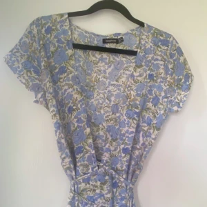 Blommig långklänning från Boohoo - Långklänning från Boohoo med blått och grönt blommönster på vit botten. Klänningen har korta ärmar, v-ringning och ett knytband i midjan för en snygg siluett. Perfekt för sommarens alla tillfällen.