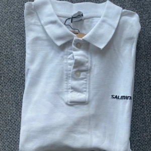 Vit pikétröja från Salming - Klassisk vit pikétröja från Salming med broderad logga på bröstet. Tröjan har krage, knäppning med tre knappar och korta ärmar. 