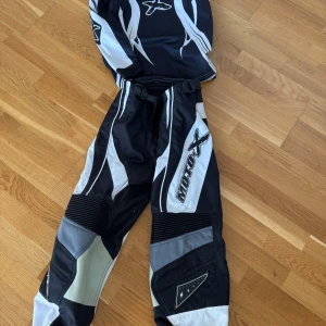 Svarta och vita motocrossbyxor Moto X - Säljer ett par svarta och vita motocrossbyxor från Moto X med coola grafiska detaljer och förstärkta partier vid knäna. Byxorna har elastisk midja och är perfekta för dig som gillar fart och action. Crosskläderna är i ett bra skick och inga hål eller skador på de!