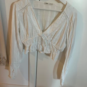 Vit croppad blus från Pull&Bear - Säljer en vit croppad blus från Pull&Bear med långa ärmar och v-ringning. Blusen har volangdetaljer och fina spetsinslag längs sömmarna. Perfekt för vår och sommar, superfin till både jeans och kjol!