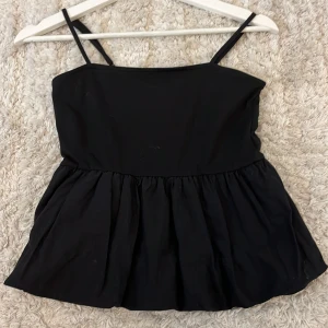 Svart volangtop från H&M Divided XS - Supersöt svart topp från H&M Divided i storlek XS. Modellen har smala axelband och en volangig, utställd nederdel som ger en luftig känsla. Perfekt att styla med jeans eller kjol för en trendig look.