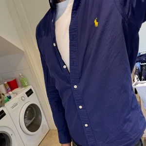 Blå Polo Ralph Lauren Skjorta - Snygg blå skjorta från Polo Ralph Lauren i slim fit-modell, storlek M. Skjortan har klassisk krage, vita knappar och den ikoniska gula loggan broderad på bröstet. Tillverkad i 100% bomull.
