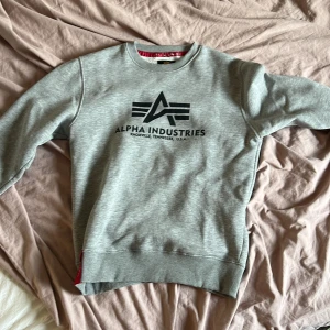 Grå sweatshirt från Alpha Industries - Säljer en grå sweatshirt från Alpha Industries med svart logga framtill. Tröjan har rund halsringning, långa ärmar och ribbade muddar. Detalj med röd etikett vid ärmslut. Perfekt för en avslappnad stil.
