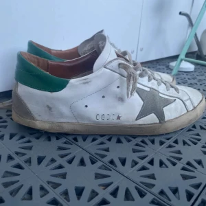Golden Goose Super-Star sneakers med grön häl - Säljer ett par vita Golden Goose Super-Star sneakers med grön detalj på hälen och grå stjärna på sidan. Skorna har snörning och platt sula, samt klassisk rund tå. Perfekta för dig som gillar en sportig och trendig look. Du får med dustbag å kvitto köpta från Carl of Carl