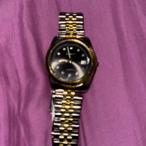 Klassisk herrklocka med silver och guld ditaler  - Stilren Casio herrklocka med svart urtavla och datumvisning. Armbandet är i rostfritt stål med guld- och silverfärgade länkar. Boetten har gulddetaljer och en räfflad kant. Perfekt accessoar för dig som gillar klassisk design.