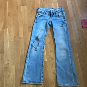 Ljusblå bootcut jeans med slitningar - Säljer ett par ljusblå jeans med bootcut passform och tydliga slitningar på ena benet och vid fickan. Jeansen har klassisk femficksdesign och normalhög midja. Perfekta för en avslappnad och trendig look. Pris kan diskuteras 💕⭐️