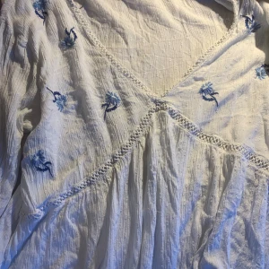 Vit blommig klänning från Flash Woman - Söt vit blus från Flash Woman i storlek XL. Blusen har blå broderade blommor, v-ringning och spetsdetaljer. Tillverkad i ett lätt bomullstyg med struktur och små prickar i tyget. Perfekt för varma dagar! Obs! Blusen har 3 små hål som går enkelt att laga!! 