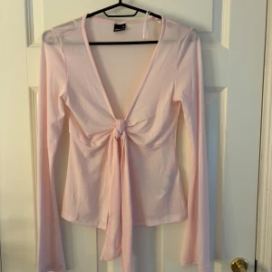 Ljusrosa omlottopp från Gina Tricot - Supersöt ljusrosa omlottopp från Gina Tricot med lång ärm och djup v-ringning. Toppen har knyt framtill som ger en snygg detalj och är gjord i mjuk bomullsmix. Perfekt för dig som gillar en feminin och trendig look.