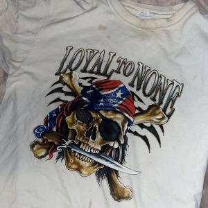 Vit t-shirt med pirattryck från Anvil - Säljer en vit t-shirt från Anvil i 100% bomull med stort tryck av en dödskalle med bandana och texten 'Loyal to None'. T-shirten har korta ärmar och en klassisk passform. Perfekt för dig som gillar edgy och unika motiv! Den har några små fläckar