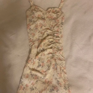 Blommig ljus kortklänning med rynk - Supersöt kortklänning med smala axelband och rynk framtill. Ljus botten med blommigt mönster i rosa och grå toner. Sitter figurnära och har en feminin, romantisk vibe. Perfekt för sommarens alla tillfällen! Använd en gång. 