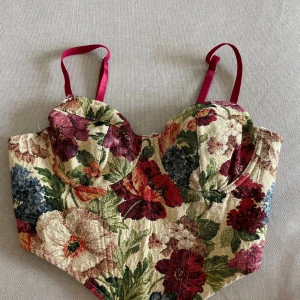 Blommig korsettopp med broderad känsla - Säljer en unik korsettopp med färgglatt blommönster i rött, grönt, blått och beige. Toppen har vadderade kupor, justerbara axelband i cerise och en brokadliknande struktur. Perfekt för dig som vill sticka ut med en vintageinspirerad look!