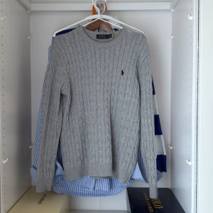 Grå kabelstickad tröja från Polo Ralph Lauren - Säljer en klassisk grå kabelstickad tröja från Polo Ralph Lauren. Tröjan har rund halsringning, lång ärm och den ikoniska broderade loggan på bröstet. Perfekt för lager-på-lager och enkel att matcha med det mesta.