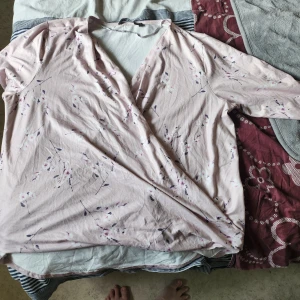 Ljusrosa omlottblus med blommigt mönster - Söt ljusrosa omlottblus med blommigt mönster i lila och vitt. Blusen har v-ringning och långa ärmar, perfekt för dig som gillar en romantisk och feminin stil. Tunn och mjuk i materialet, passar bra till både jeans och kjol.