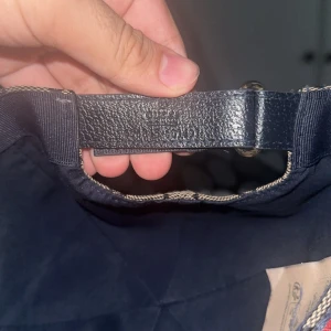 Gucci keps med GG-mönster och röd detalj - Snygg keps från Gucci i beige med klassiskt GG-monogram över hela kepsen. Baksidan har en justerbar rem i svart skinn och en ikonisk röd och marinblå rand. Kepsen är tillverkad i bomullsmix och har en böjd skärm.