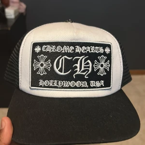 Chrome Hearts truckerkeps svart/vit - Snygg truckerkeps från Chrome Hearts i svart och vitt med broderad logga och text på framsidan. Kepsen har mesh-bakstycke, justerbar snapback och metallknapp med gravyr på toppen. Perfekt statement-piece för streetwear.