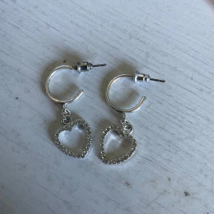 Silverörhängen med hjärtan och stenar - Snygga örhängen i silverfärg med öppna hjärtan som hänger i små ringar. Hjärtana är dekorerade med glittrande stenar som ger extra bling. Perfekta för dig som gillar söta detaljer och vill sticka ut lite extra.