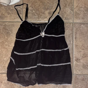 Svart spetsig linnetopp med rosett - Svart linnetopp från H&M med tunna axelband och transparent mesh. Toppen har spetsdetaljer, ljusgrå spetskant och en söt rosett framtill. Lätt och luftig känsla, perfekt för lager-på-lager eller som statement-piece.