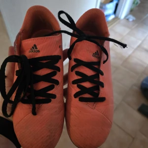 Adidas orange fotbollsskor strl 33 - Säljer ett par orangea fotbollsskor från Adidas med svarta snören och dobbar för gräsplan. Skorna har en sportig look och är tillverkade i syntetmaterial med textilfoder. Perfekta för dig som vill sticka ut på planen!