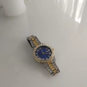 Diamant  klocka med blå urtavla - Säljer en riktigt blingad klocka med blå urtavla, romerska siffror och datumvisning. Armbandet är i silver och guld med massor av glittrande stenar. Klockan har rund form och är perfekt för dig som gillar att sticka ut med accessoarer.