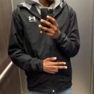 Svart vindjacka från Under Armour - Säljer en svart vindjacka från Under Armour med dragkedja och huva. Jackan har en sportig look och logga på bröstet. Perfekt för utomhusaktiviteter och enkel att matcha med olika outfits.