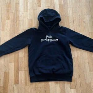 Svart hoodie från Peak Performance - Säljer en svart hoodie från Peak Performance med vit broderad logga på bröstet. Tröjan har huva och en stor magficka framtill. Perfekt för dig som gillar en klassisk och stilren look.