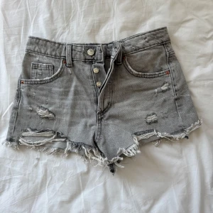 Grå jeansshorts med slitningar från Zara - Gråa jeansshorts från Zara med råa, fransiga kanter och slitna detaljer. Klassisk femficksmodell med hög midja och knappgylf. Perfekta för en avslappnad och trendig look.