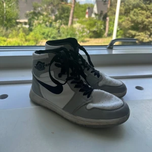 Nike Air Jordans i vitt och grått - Säljer ett par klassiska skor i vitt och grått med svarta detaljer och snörning. Skorna har hög modell och svart swoosh på sidan. Perfekta för dig som gillar streetwear och vill ha en ikonisk sneaker i garderoben. Ej äkta 