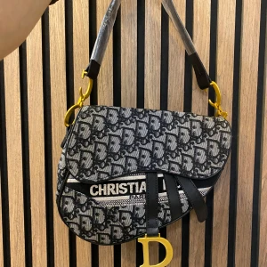  Saddle Bag axelväska - Säljer en ikonisk  Saddle Bag axelväska i svart och grått med tydligt monogram över hela väskan. Väskan har guldfärgade metalldetaljer, bred axelrem med logga och en stor D-charm framtill. Materialet är canvas med läderdetaljer.