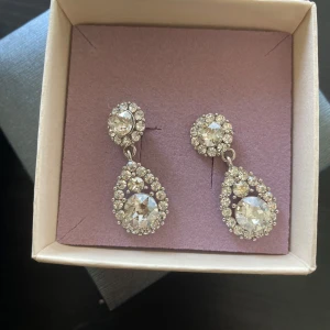 Glittriga örhängen från Lily and Rose - Säljer ett par örhängen från Lily and Rose. Örhängena är silverfärgade med stora stenar i droppform och runda stenar upptill, omgivna av mindre kristaller. Använt en gång till studentbal, som nya.