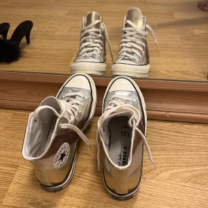 Silvriga, guld  och bronsfärgade Converse Chuck Taylor All Star - Säljer ett par höga Converse Chuck Taylor All Star med metallic silvriga och bronsfärgade paneler. Skorna har vit sula, vita snören och klassisk rund tå. Perfekta för dig som vill sticka ut med en unik look.
