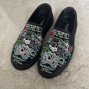 Svarta slip-on sneakers från Kenzo med broderad tiger - Säljer ett par svarta slip-on sneakers från Kenzo med färgglad broderad tiger och textdetaljer på ovandelen. Skorna har rund tå och platt sula, perfekt för dig som vill sticka ut med din stil.