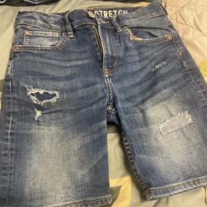 Blå slitna jeansshorts med stretch - Snygga blå jeansshorts med slitningar och lagade detaljer. Modellen har klassisk femficksdesign, dragkedja och knapp framtill samt stretchigt material för extra komfort. Perfekta för en avslappnad och trendig look.