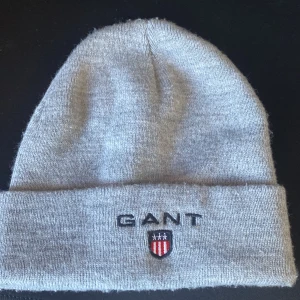 Grå stickad mössa från GANT - Snygg grå stickad mössa från GANT med broderad logotyp och sköld framtill. Klassisk modell med uppvikt kant, perfekt för kyliga dagar. Enkel och stilren design som passar till det mesta.