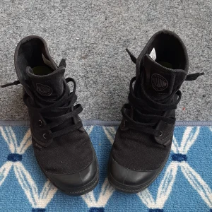 Svarta Palladium boots i canvas - Säljer ett par svarta boots från Palladium i slitstark canvas med grov sula och rund tå. Skorna har snörning och diskret logga på sidan och plösen. Perfekta för dig som gillar en urban och avslappnad stil.