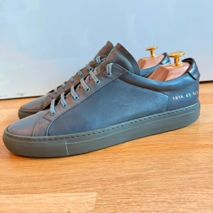 Gråa sneakers i skinn från Common Projects - Snygga minimalistiska sneakers från Common Projects i grått skinn med matchande grå snörning och diskret guldfärgad numrering på hälen. Klassisk rund tå och platt sula ger en clean look som passar till allt. Perfekta för dig som gillar stilren design. Nypris ligger på över 4000 säljer de för billigt för behöver få bort de snabbt och köpa nya skor då de var för stora för mig