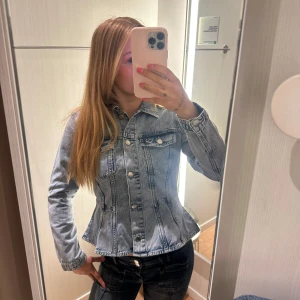 Ljusblå jeansjacka med knappar - Säljer en snygg ljusblå jeansjacka med klassisk krage och silverfärgade knappar framtill. Jackan har två bröstfickor med lock och markerad midja som gör den figursydd. Bra skick o enbart använt 2 gånger! 