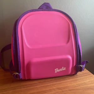 Rosa Barbie ryggsäck - Säljer en söt rosa och lila ryggsäck från Barbie. Perfekt för dig som gillar färgglada accessoarer och vill sticka ut lite extra. Är ungefär 28 cm hög och ca 25-30 cm bred.  Har två små vita märken på framsidan.