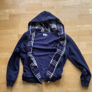 Mörkblå hoodie från Burberry med rutigt foder - Säljer en mörkblå hoodie från Burberry med klassiskt rutigt foder i huvan och längs dragkedjan. Tröjan har dragkedja framtill, långa ärmar och broderad logga på bröstet. Perfekt för dig som gillar stilrena detaljer och lyxiga märken.