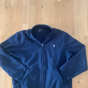 Mörkblå ziptröja från Polo Ralph Lauren - Säljer en mörkblå ziptröja från Polo Ralph Lauren med klassisk vit logga på bröstet. Tröjan har dragkedja framtill, ståkrage och två fickor. Perfekt för en stilren och avslappnad look.