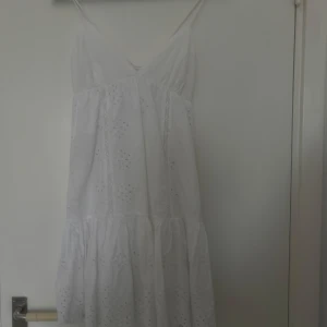 Vit somrig klänning  - vit klänning med smala axelband och v-ringning. Klänningen har broderade blommor, volang nertill och snörning i ryggen. Perfekt för varma dagar och midsommarfirande.