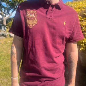 Vinröd pikétröja från Polo Ralph Lauren - Säljer en vinröd pikétröja från Polo Ralph Lauren i modellen Custom Slim Fit. Tröjan har korta ärmar, klassisk krage och knappar framtill. Broderad gul logga på bröstet och tryck på vänster sida. Perfekt för en stilren look.