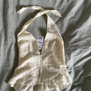 Ljusgul halterneck topp med knappar - Supersöt ljusgul halterneck topp från Stradivarius med knappar framtill och lätt v-ringning. Toppen har ett subtilt randigt mönster och knyts i nacken. 
