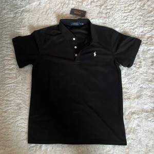 Polo Ralph Lauren Tröja - Klassisk svart pikétröja från Polo Ralph Lauren med vit broderad logga på bröstet. Tröjan har korta ärmar, krage och tre vita knappar framtill. Tillverkad i mjuk bomull som känns skön mot huden. Perfekt för en clean och stilren look. Helt ny och oanvänd.