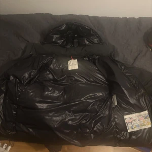  Moncler mongenevre  - Säljer en svart Moncler dunjacka med huva och klassisk logga på ärmen. Jackan har blanka detaljer, dragkedjor och quiltad design. Perfekt för kalla dagar och riktigt snygg till streetwear. Fodrad och med flera fickor för mobilen och annat viktigt. Pris ej fast 