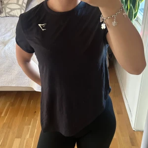 Svart t-shirt från Gymshark - En svart t-shirt från Gymshark med rund hals och korta ärmar. T-shirten har en liten vit Gymshark-logga på bröstet och är tillverkad i mjuk bomull. Perfekt för träning eller en avslappnad look. Slits på sidan som antigen går att ha som den är eller knyta☺️SÄLJER LIKADAN I VIT