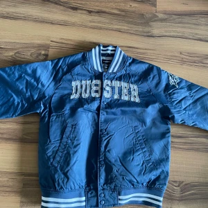 Blå bomberjacka från DUBSTER - Säljer en blå bomberjacka från Dubster med vita ränder på krage, ärmslut och midja. Jackan har tryckknappar, två fickor och broderad logga på bröstet. Perfekt för dig som gillar sportig stil!